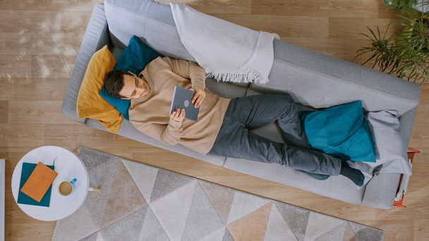 Young Man in Brown Jumper and Grey Jeans is Lying Down on a Sofa, Using a Tablet. He is Happy and Smiles. Cozy Living Room with Modern Interior with Plants, Table and Wooden Floor. Top View. - Photo, Image