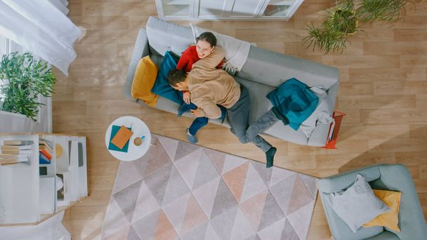 Young Beautiful Couple is Fooling and Partying Around. Running Across the Room and Lying Down on Sofa. Cozy Living Room with Modern Interior with Plants, Table and Wooden Floor. Top View. - 写真・画像