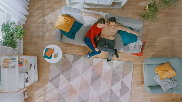 Young Beautiful Couple Sitting on a Couch with Pillows and Watching TV. They are Excited and Smile. Cozy Living Room with Modern Interior with Plants, Table and Wooden Floor. Top View. - Foto, afbeelding