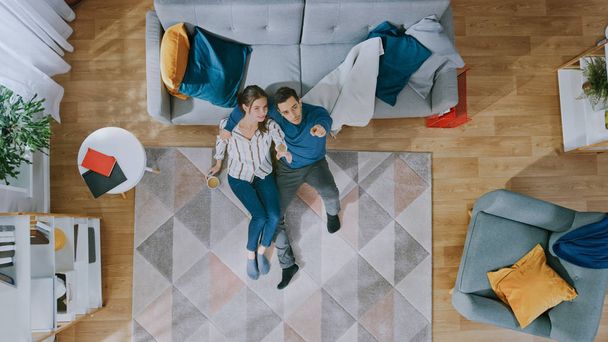 Couple is Sitting on a Floor, Talking and Laughing. Man Gives Girl a Hug. They Look Above. Living Room with Modern Interior with Carpet, Sofa, Chair, Table, Shelf, Plant and Wooden Floor. Top View. - Zdjęcie, obraz