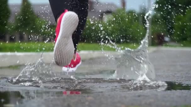 Spor ayakkabılı bir koşucunun bacakları. Spor kadını dışarıda koşuyor, çamurlu su birikintisine giriyor. Yağmurda koşan, su sıçratan tek koşucu. Arka görüş. Yavaş çekim - Video, Çekim
