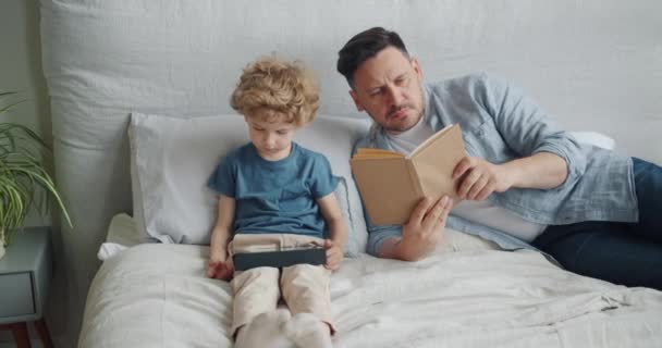 Serious guy reading book in bed while small child watching cartoons on tablet - Záběry, video