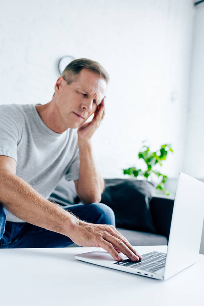 hombre guapo en camiseta con dolor de cabeza usando el ordenador portátil en el apartamento
  - Foto, imagen