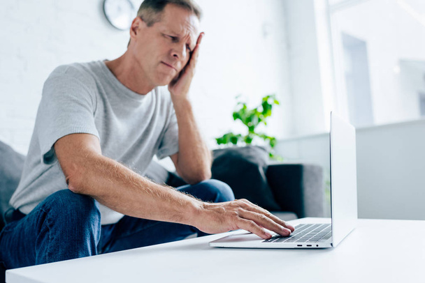 hombre guapo en camiseta con dolor de cabeza usando el ordenador portátil en el apartamento
  - Foto, imagen