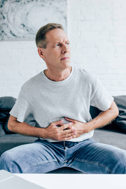 handsome man in t-shirt with stomachache looking away in apartment  - Photo, image