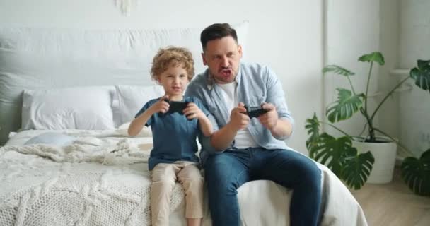 Middle-aged guy playing video game with happy child sitting on bed at home - Materiał filmowy, wideo