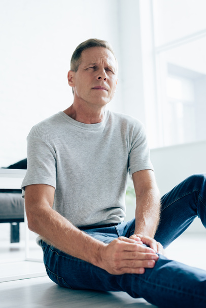 pohledný muž v tričku cítí bolest v koleni v bytě  - Fotografie, Obrázek