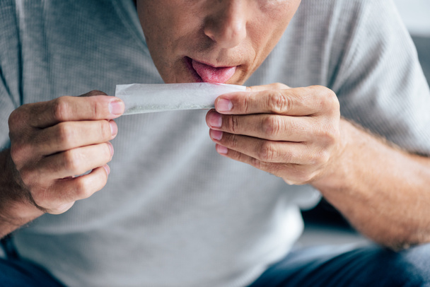 cropped view of man in t-shirt licking paper for blunt  - Фото, изображение