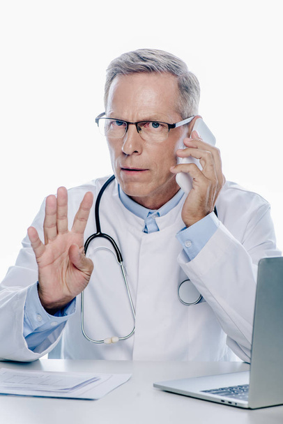 médico bonito em casaco branco falando no smartphone e mostrando gesto isolado no branco
 - Foto, Imagem