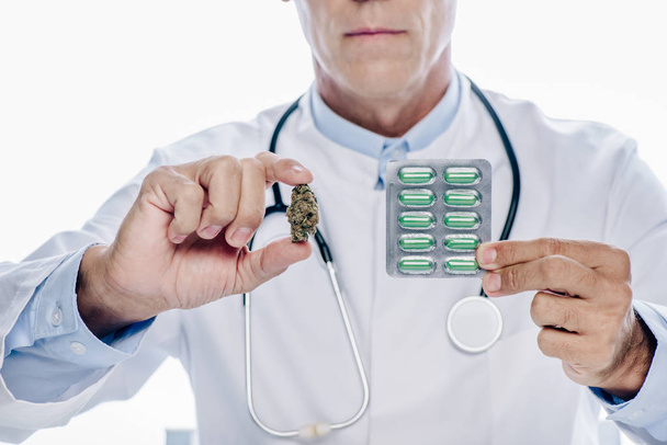 cropped view of doctor in white coat holding medical marijuana and pills isolated on white - Fotografie, Obrázek