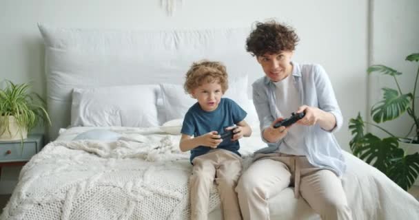 Mother and son playing video game sitting on bed in bedroom at home having fun - Video