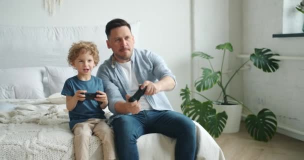 Cute kid playing video game with father holding joysticks having fun in bed - Video
