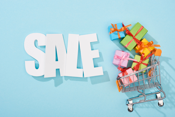 top view of white sale lettering and colorful gift boxes in shopping cart on blue background - Fotoğraf, Görsel
