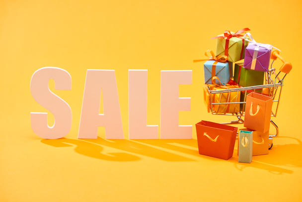 witte verkoop belettering met boodschappentassen en feestelijke geschenken in winkelwagen op heldere oranje achtergrond - Foto, afbeelding