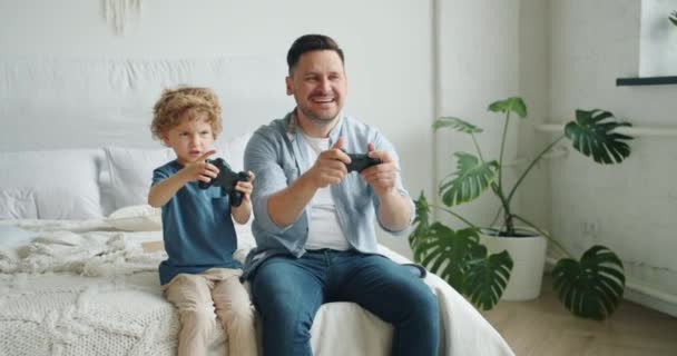 Slow motion of man and boy playing video game having fun sitting in bed at home - Video, Çekim