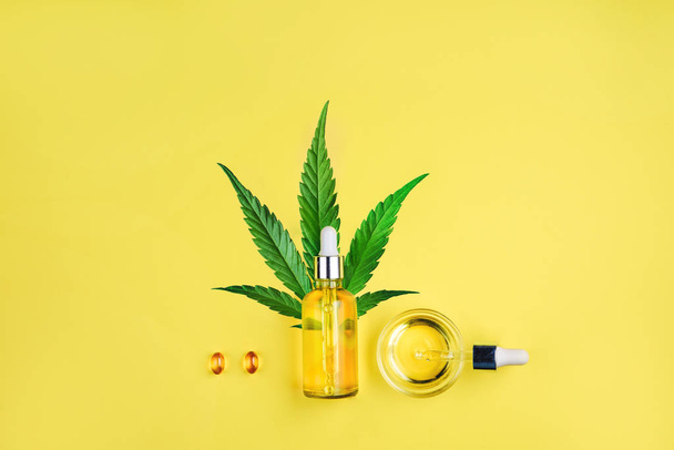 Bottle with CBD oil, pipette, capsules and cannabis leaf on a yellow background. Minimalism, flat lay. - Photo, Image