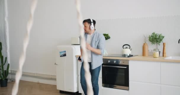 Handsome guy dancing in kitchen wearing headphones holding smartphone - Séquence, vidéo