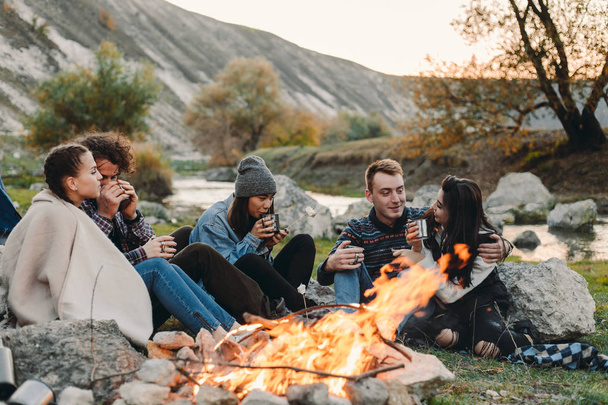 Sonriendo amigos carismáticos diferentes razas en el camping, sentado al lado de una hoguera y beber algunas bebidas calientes de copas metálicas y pasar un tiempo increíble juntos
. - Foto, imagen