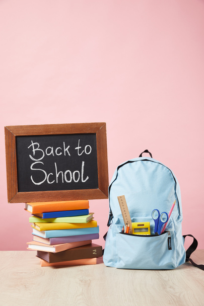 blauwe rugzak met benodigdheden in zak in de buurt van schoolbord met terug naar school belettering op boeken geïsoleerd op roze - Foto, afbeelding