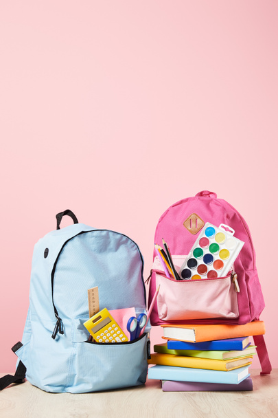 mochilas azules y rosas con útiles escolares cerca de pila de libros aislados en rosa
 - Foto, Imagen