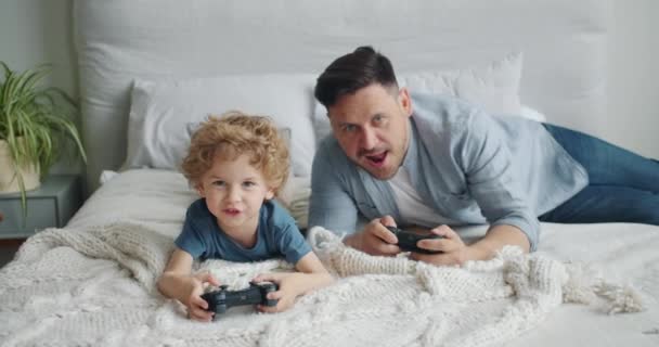 Ojciec i dziecko gra wideo na łóżku w domu naciskając przyciski na joysticku - Materiał filmowy, wideo
