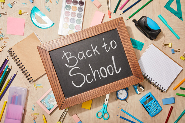 Top zicht van verschillende schoolbenodigdheden en schoolbord met terug naar school belettering op houten bureau - Foto, afbeelding