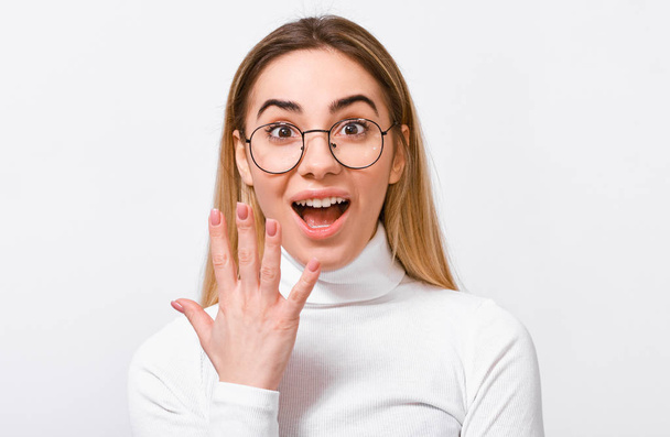 Πορτρέτο του ενθουσιασμένος νεαρή ξανθιά γυναίκα φορώντας λευκή μπλούζα και στρογγυλά μοντέρνα διαφανή γυαλιά, δείχνοντας δαχτυλίδι αρραβώνων στο δάχτυλό της, ποζάρει πάνω από λευκό φόντο στούντιο. - Φωτογραφία, εικόνα