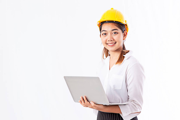 Piękna Młoda Azjatycka kobieta ubrana w żółty kask ochronny. Architekt kobieta za pomocą laptopa i uśmiechając się stojąc w jej miejscu pracy z białym tle i przestrzeni kopiowania. - Zdjęcie, obraz