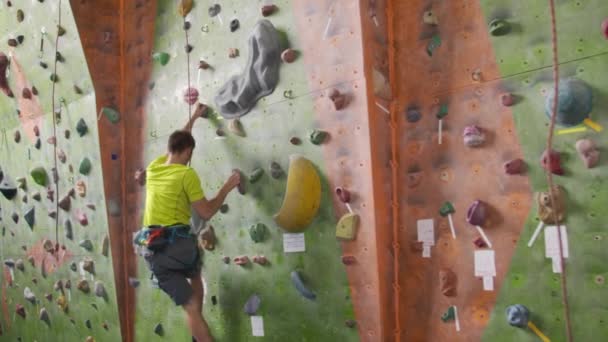Male climber climbs the cliff wall in the room reaching and gripping hold. A professional concentrated climber moves up the wall, the camera rises and lays down his movements. Boom follow camera - Footage, Video