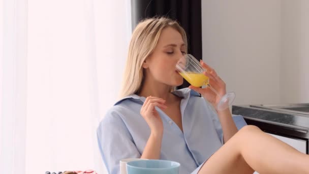Tracking out colpo di bella donna bionda sorridente in camicia maschile mangiare croissant godendo il gusto e bere succo d'arancia sulla cucina moderna
 - Filmati, video