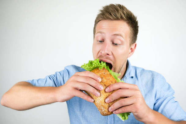 young handsome hungry man eats fresh sandwich with salad leaf on isolated white background - Фото, изображение