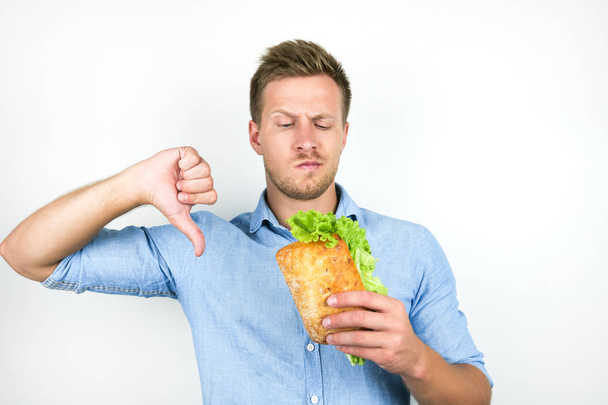 young handsome hungry man looks hungry holding fresh sandwich with salad leaf showing dislike sign on isolated white background - Фото, изображение