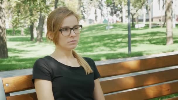Η όμορφη γυναίκα με τα γυαλιά κοιτάζει την κάμερα και χασμουριέται. Ελκυστικό κορίτσι κάθεται στον πάγκο στο πάρκο της πόλης και χασμουριέται - Πλάνα, βίντεο