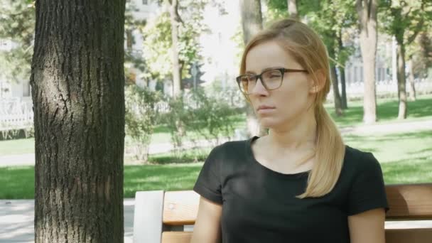 Hübsche Frau mit Brille blickt in die Kamera und gähnt. attraktives Mädchen sitzt auf Bank im Stadtpark und gähnt - Filmmaterial, Video