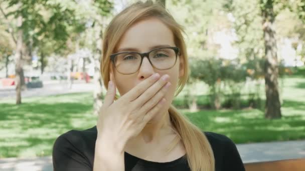 Pretty woman in glasses looks at the camera and yawns. Attractive girl sits on the bench in city park and yawns - Footage, Video