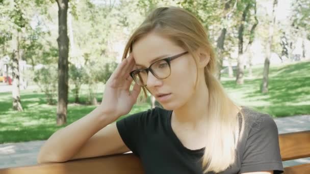 Молода жінка в окуляри з головним болем сидить на лавці в міському парку. Кавказька дівчина відчувати себе нещасливою і з мігрені торкаючись її храмів. Страшна мігрень. - Кадри, відео