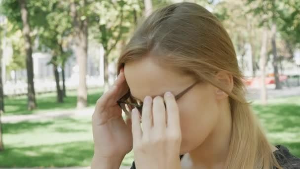 Молода жінка в окуляри з головним болем сидить на лавці в міському парку. Кавказька дівчина відчувати себе нещасливою і з мігрені торкаючись її храмів. Страшна мігрень. - Кадри, відео