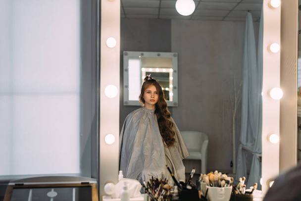 Bir güzellik stüdyosunda bir sandalyede oturan kıvrılmış saçlı kız. Yansımayı aynada görüntüleme, - Fotoğraf, Görsel