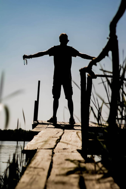 Cheerful guy stands on the pier near the river and looks at the horizon one - Φωτογραφία, εικόνα
