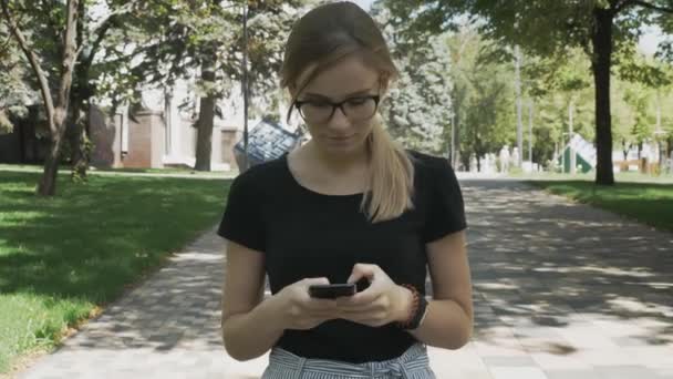 Porträt einer jungen schönen Frau, die per App auf dem Smartphone lächelt und auf dem Handy SMS schreibt. Kaukasische Frau mit Brille spaziert im Stadtpark. - Filmmaterial, Video
