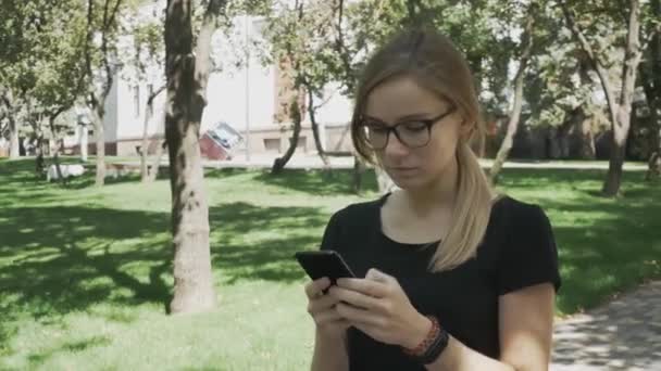 Perplexed caucasian woman in eyeglasses walking checking smart phone content and then stops looking at camera in a park - Footage, Video