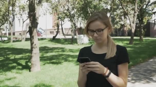 Perplexed caucasian woman in eyeglasses walking checking smart phone content and then stops looking at camera in a park - Video