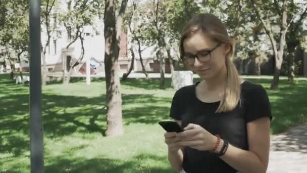 Pretty young caucasian woman in eyeglasses walking towards camera, checking phone and celebrating good news in a park - Imágenes, Vídeo