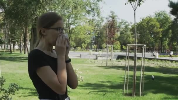 young ill woman in eyeglasses walking sneezing in a city park in summer day - Materiał filmowy, wideo