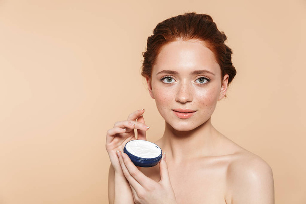 Amazing young redhead woman posing isolated over beige wall background take care of her skin with cream. - Фото, зображення