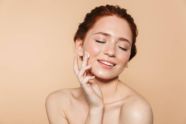 Amazing young redhead woman posing isolated over beige wall background take care of her skin with cream. - Zdjęcie, obraz