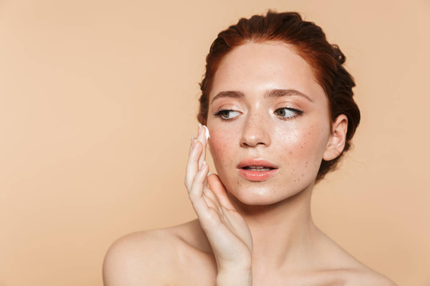 Amazing young redhead woman posing isolated over beige wall background take care of her skin with cream. - Фото, изображение