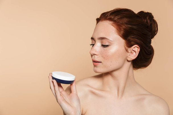 Amazing young redhead woman posing isolated over beige wall background take care of her skin with cream. - Valokuva, kuva