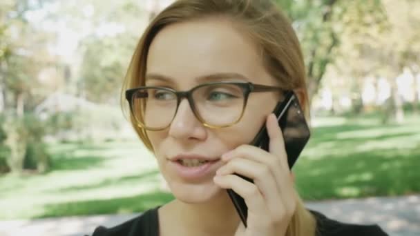 Χαρούμενη νεαρά καυκάσια γυναίκα με γυαλιά που μιλούν στο μαύρο κινητό τηλέφωνο ενώ κάθονται στο παγκάκι στο πάρκο της πόλης την καλοκαιρινή μέρα. Κλείστε πορτραίτα.  - Πλάνα, βίντεο