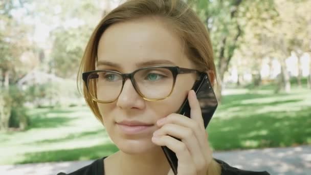 Счастливая молодая белая женщина в очках разговаривает по черному телефону, сидя на скамейке в городском парке в летний день. Крупный план портретов
.  - Кадры, видео